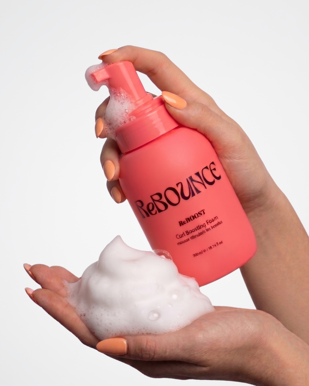 Hand holding a ReBOOST pink bottle of ReBOUNCE curl boosting foam product with foam on a white background