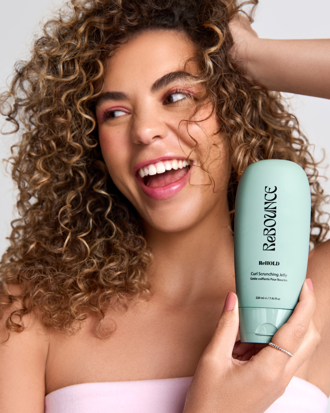 Woman with curly hair holding a bottle of ReBOUNCE product against a neutral background